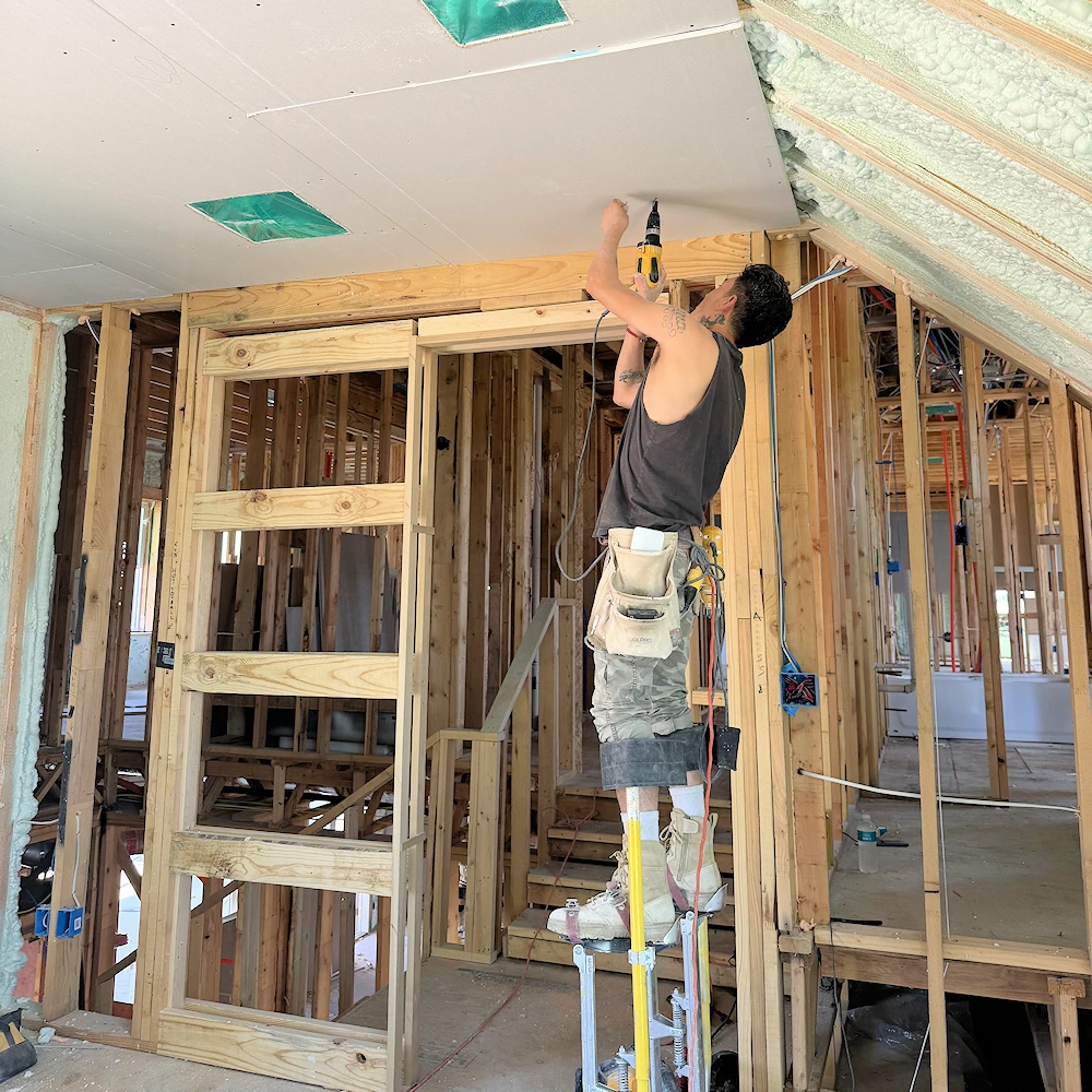 Worker on stilts hanging drywall on a ceiling.